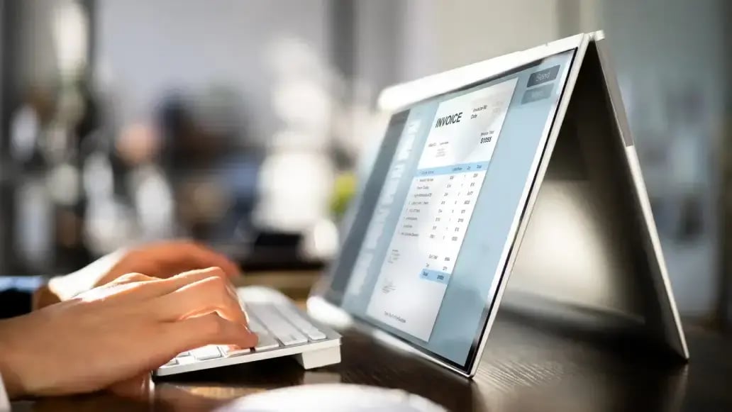 Person types on keyboard while an e-invoicing invoice screen is visible on a laptop in tent position on a desk.